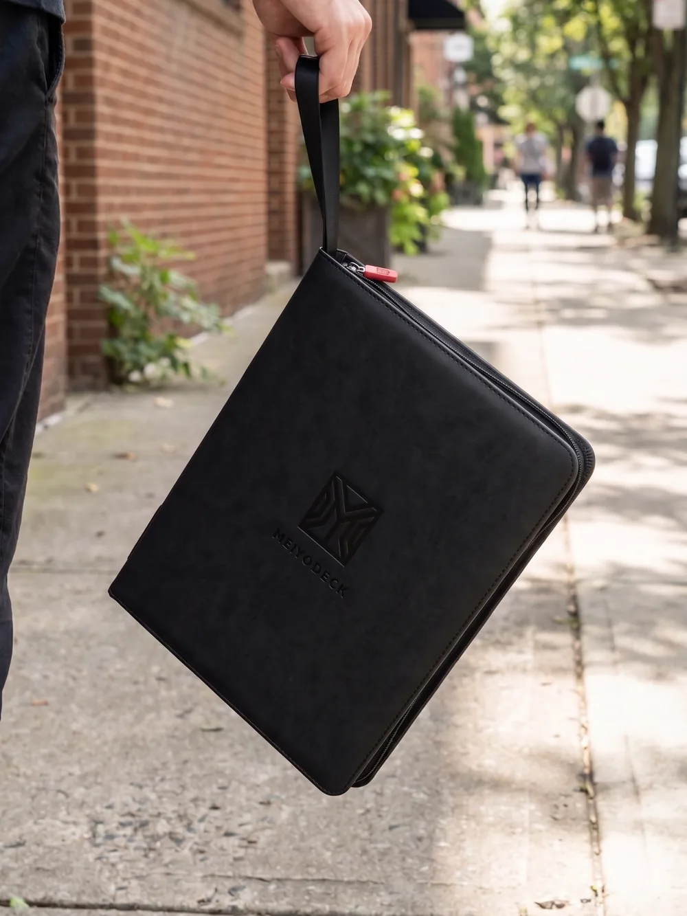 A hand carrying the black Meiyodeck top loader binder by its strap on a city sidewalk